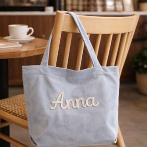 Puede incluir: Bolso tote de pana azul claro con el nombre "Anna" bordado en escritura color crema. El bolso está colgado de una silla de madera en un café. El bolso tiene dos asas y forma rectangular.