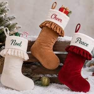 May include: Three Christmas stockings with white cuffs and ruffled trim. The cream stocking features the green embroidered text 'Noel', the tan stocking has the red embroidered text 'Cheer', and the red stocking has the green embroidered text 'Peace'.