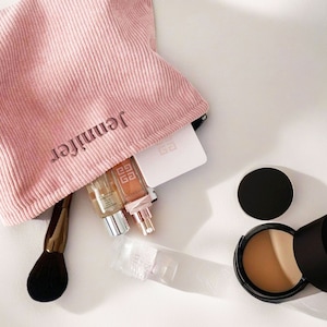 Op de afbeelding: Een roze corduroy make-up tas met de naam "Jennifer" erop geborduurd. De tas is open en bevat verschillende make-up producten, waaronder een foundation compact, een fles serum en een kwast.