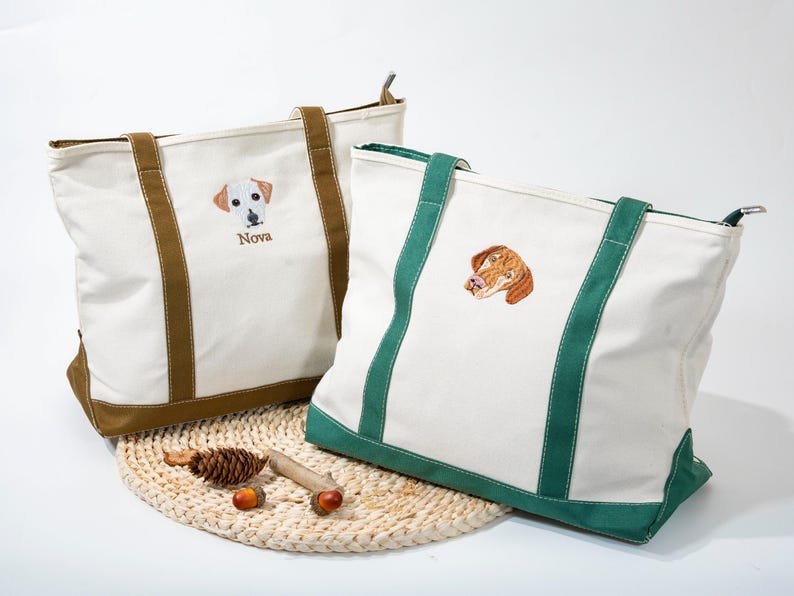 May include: Two white canvas tote bags with embroidered dog portraits. One bag has brown accents and the name "Nova" embroidered below the dog. The other bag has green accents. Both bags have sturdy handles and a zippered top.