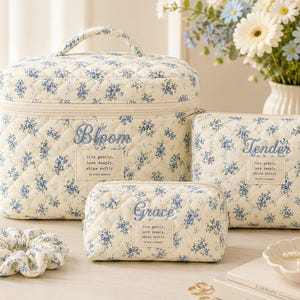 Puede incluir: Un conjunto de tres neceseres acolchados en color crema con estampado floral azul. Cada bolsa presenta texto bordado que incluye Bloom, Tender y Grace, junto con una etiqueta rectangular que dice live gently, love deeply, shine softly, in every moment.