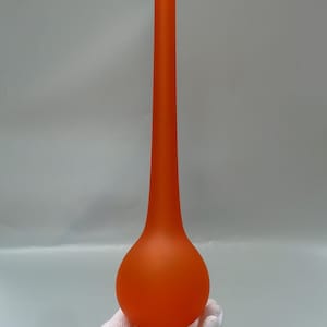 May include: A tall, orange glass vase with a bulbous base and a long, slender neck. The vase has a matte finish and sits on a small, frosted glass base. The overall shape is elegant and modern.