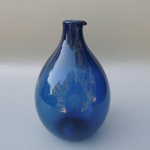 May include: A cobalt blue glass vase with a rounded, teardrop shape and a small spout. The vase is smooth and reflective, and the blue color is deep and rich.