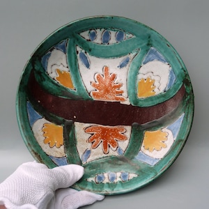 May include: A round ceramic plate with a green background and a colorful geometric design. The design features orange, yellow, blue, and white accents.