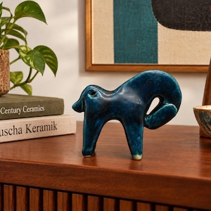May include: A small, stylized ceramic animal figurine with a glossy, deep blue glaze. The sculpture features a curved back, four short legs, and a rounded tail, standing upright on a smooth, polished wooden surface.