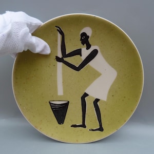 May include: A round, speckled, light green plate with a black and white illustration of a person grinding. The person is wearing a white dress and is holding a long white pole over a black container.