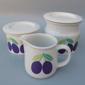 May include: A set of three white ceramic kitchenware pieces with a plum design. The set includes a creamer, a sugar bowl with a lid, and a jar with a lid. Each piece features a stylized plum design with green leaves.