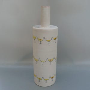 May include: A tall, cylindrical ceramic vase with a narrow neck and a wide base. The vase is white with a repeating pattern of yellow cocktail glasses outlined in black. The rim of the neck is pink.