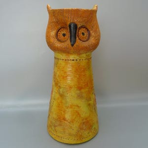 May include: A tall, ceramic owl vase with a yellow and orange glaze. The owl's eyes are black and white, and its beak is black.
