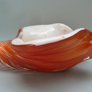 May include: A decorative glass bowl with a unique shape and color. The bowl features a vibrant orange exterior with a swirling pattern and a white interior. The glass has a glossy finish, adding to its visual appeal.