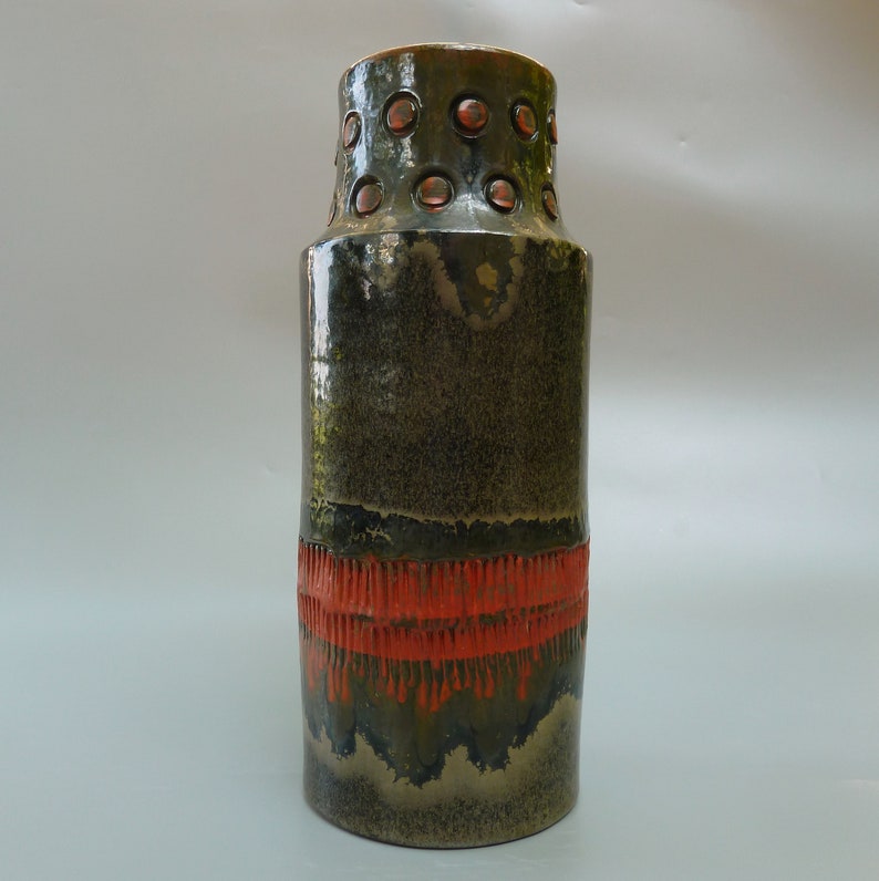 May include: A tall, cylindrical ceramic vase with a dark green glaze and a band of orange glaze around the middle. The vase has a textured surface and a series of small, round holes around the top.