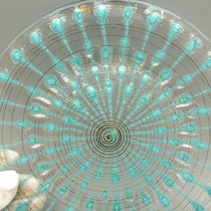 May include: A clear glass plate featuring a central gold spiral design surrounded by radiating turquoise teardrop shapes and thin gold circular lines. The glass surface displays a repeating pattern of teal accents and metallic gold detailing.