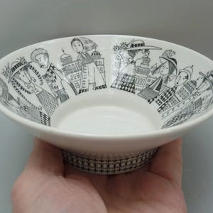 May include: White ceramic bowl with a black and white illustration of people and buildings around the rim. The bowl has a decorative base with a repeating pattern. The bowl is held in a hand.