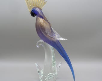 Peut inclure: Une sculpture en verre représentant un perroquet avec une crête jaune, un corps bleu et un perchoir transparent. Le corps de l'oiseau présente un dégradé de couleurs, passant du marron clair au bleu profond. La sculpture est détaillée et élégante.
