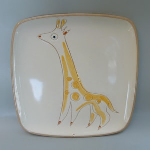 May include: A square ceramic plate with a hand-painted design of a yellow giraffe with brown spots. The giraffe is facing left and has a long neck and legs.