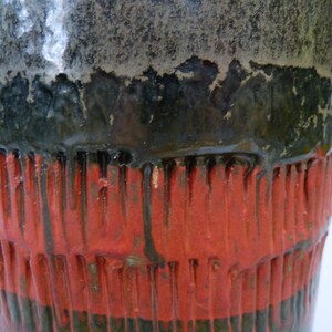 May include: Close-up of a ceramic vase with a black, green, and red glaze. The vase has a textured surface with vertical lines.