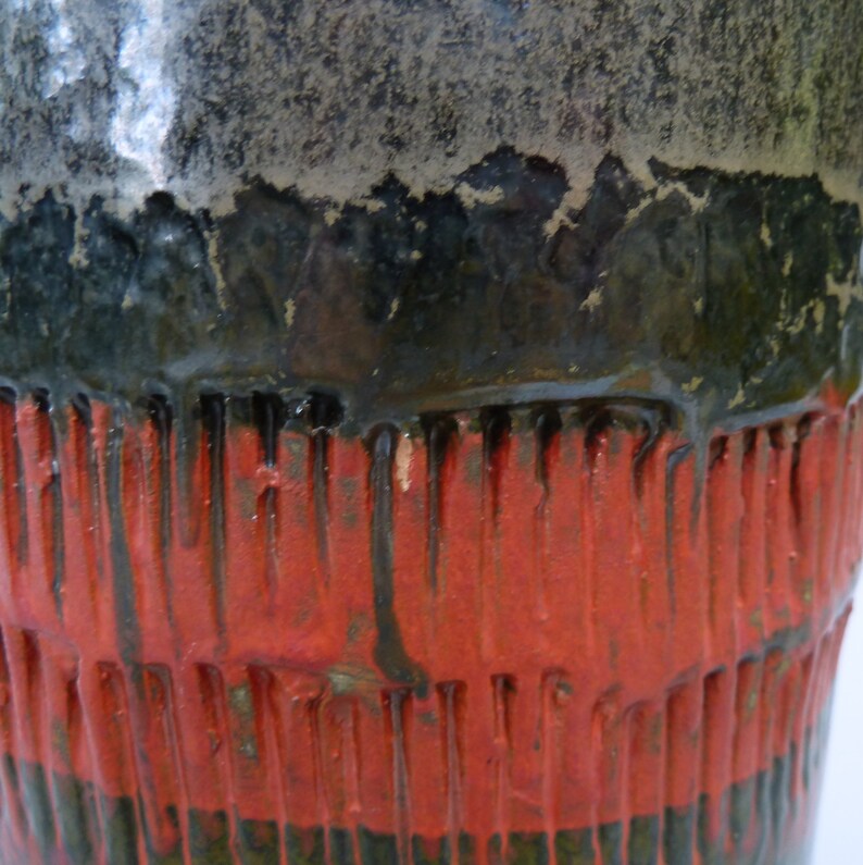 May include: Close-up of a ceramic vase with a black, green, and red glaze. The vase has a textured surface with vertical lines.