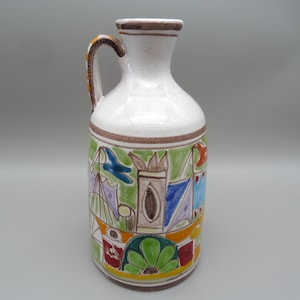 May include: A white ceramic pitcher with a single handle and a narrow neck. The body features a hand-painted, colorful design depicting stylized architectural structures, geometric shapes, and floral motifs in shades of green, blue, yellow, and red.