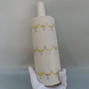 May include: A white ceramic bottle with a cylindrical body and a long, narrow neck. The bottle is decorated with rows of yellow cocktail glasses, each with a small red dot. The bottle is held by a gloved hand.