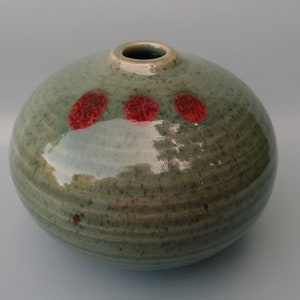 May include: A round, green ceramic vase with three red spots. The vase has a textured surface and a slightly flared opening.