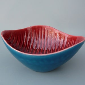 May include: A teal blue ceramic bowl with a red interior. The bowl has a wavy, leaf-like shape and a textured, ribbed surface.