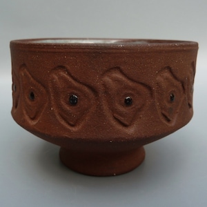 May include: A brown ceramic bowl with a footed base. The bowl has a decorative pattern of stylized flowers with black accents around the rim.