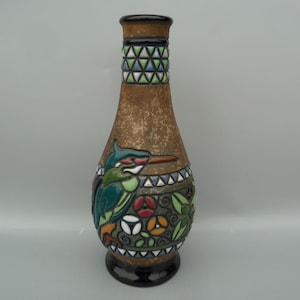 May include: A brown ceramic vase with a geometric pattern around the neck and a colorful bird design on the body. The bird is green, blue, and yellow with a long beak. The vase is decorated with flowers and leaves in shades of green, red, and orange.