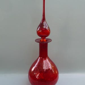 May include: A tall, red glass bottle with a long, pointed stopper. The bottle has a rounded base and a slender neck. The stopper is a teardrop shape. The glass has a textured, crackled appearance.