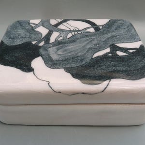 May include: A rectangular, white ceramic trinket box with a lid. The lid is decorated with a dark teal and gray abstract design. The box has rounded edges and a glossy finish, suitable for storing small items.