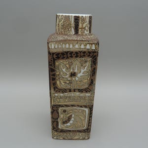 May include: A tall, rectangular ceramic vase featuring a textured surface with a brown and cream color palette. The exterior is decorated with intricate, repeating geometric patterns and stylized floral motifs arranged in stacked square panels.