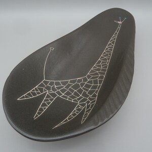 May include: A dark brown, teardrop-shaped ceramic dish featuring a stylized white line drawing of a giraffe. The giraffe has a mosaic-like pattern on its body, a small pink detail on its head, and two small blue dots representing its horns.
