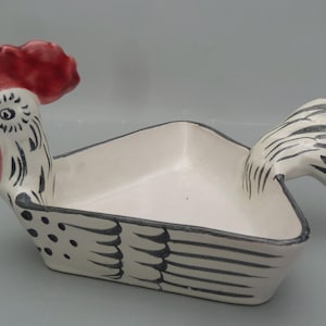 May include: A ceramic rooster-shaped serving dish. The dish is white with black and red accents. The rooster's comb and wattle are red, and the body has black detailing. The dish is triangular with a hollow center.
