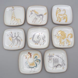 May include: Nine square ceramic plates with hand-painted animal designs. The plates feature a giraffe, zebra, tiger, camel, hippo, lion, rooster, and bull. Each plate has a white background with brown trim and colorful animal illustrations.