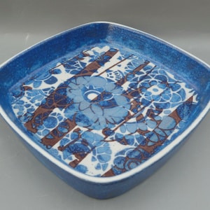May include: A square ceramic serving dish with a blue glaze and a white floral pattern in brown. The dish has a raised rim and a slightly textured surface.