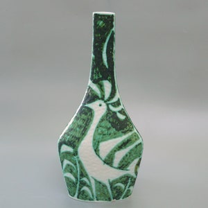 May include: A white ceramic vase with a green and white stylized bird design. The vase has a narrow neck and a wide base.