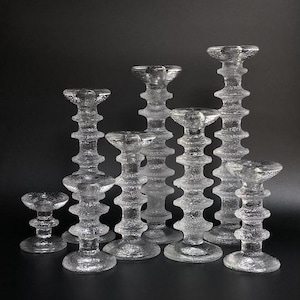 May include: A collection of clear glass candle holders of varying heights. The candle holders have a textured, ribbed design and a wide base. The tallest candle holder is approximately 10 inches tall. The candle holders are arranged on a dark background.