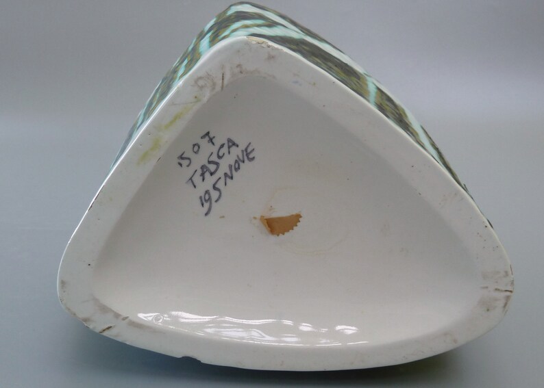 Puede incluir: La parte inferior de un jarr&oacute;n de cer&aacute;mica triangular con un patr&oacute;n abstracto verde y blanco. El jarr&oacute;n est&aacute; marcado con el texto "1507 TASCA 1957 NOVE".