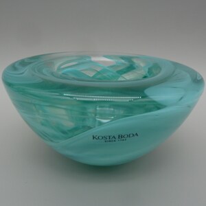 May include: A clear and turquoise glass bowl with a swirling pattern throughout the material. The exterior surface features the black printed text KOSTA BODA SINCE 1742. The bowl has a rounded, thick-walled design with a smooth, polished finish.