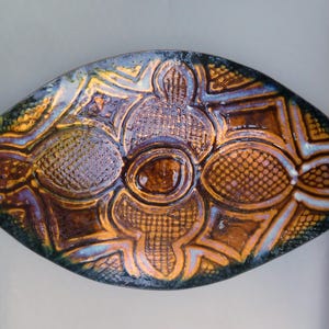 May include: An eye-shaped ceramic dish with a textured surface and intricate embossed patterns. The dish features a blend of brown, gold, and blue hues, creating a unique and artistic appearance. The design includes geometric and floral motifs.