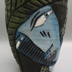 May include: A ceramic vase with a blue and green abstract design. The vase has a textured surface and is decorated with a stylized face.