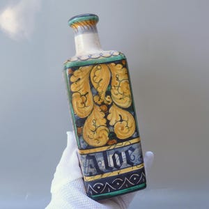 May include: A rectangular ceramic bottle with a decorative design. The bottle features a yellow and blue floral pattern with the word "Alor" in blue. The bottle's neck is white with green and yellow accents.