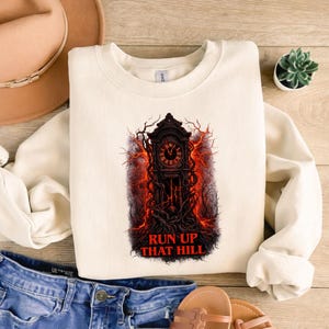 May include: Cream-colored sweatshirt featuring a graphic of a grandfather clock with red lightning and tree-like vines. The text "RUN UP THAT HILL" is printed in red below the clock. The sweatshirt is made of a soft material.