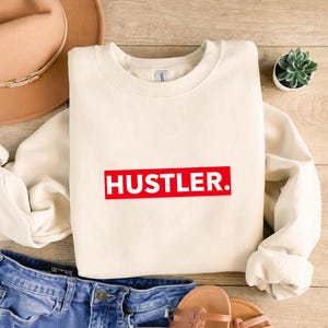 May include: Cream-colored sweatshirt with the word "HUSTLER." in bold white letters inside a red rectangle. The sweatshirt is folded on a wooden surface, with a pair of blue jeans, a hat, and sandals visible.