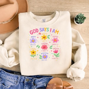 Puede incluir: Sudadera crema con el texto "GOD SAYS I AM" y gráficos florales. El diseño incluye palabras como "Brave", "Worthy" y "Beautiful", con versículos bíblicos correspondientes. La sudadera se combina con vaqueros azules y sandalias.
