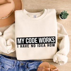 Pode incluir: Camisola creme com o texto "MY CODE WORKS I HAVE NO IDEA HOW" num retângulo preto. A camisola está dobrada sobre uma superfície de madeira, com um chapéu, calças de ganga e sandálias no fundo.