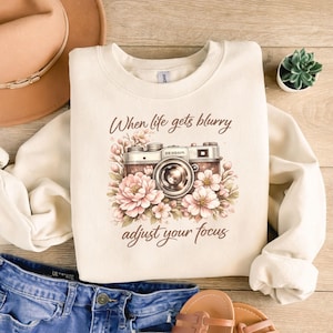 May include: Cream-colored sweatshirt with a vintage camera graphic surrounded by pink and white flowers. The text "When life gets blurry adjust your focus" is printed above and below the camera image. The sweatshirt is paired with blue jeans and sandals.