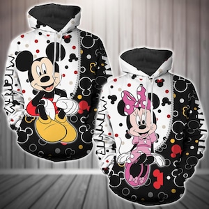 Puede incluir: Sudadera con capucha blanca y negra con un diseño colorido de Mickey Mouse y Minnie Mouse. El diseño presenta a Mickey Mouse sentado con las piernas cruzadas y a Minnie Mouse de pie con las manos en las caderas. El fondo es un patrón de lunares blancos y negros.