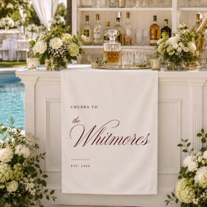 May include: A white fabric table runner draped over a bar, featuring the dark red printed text "CHEERS TO the Whitmores EST. 2026". The runner is positioned between two large floral arrangements of white roses and greenery, with a glass decanter above.