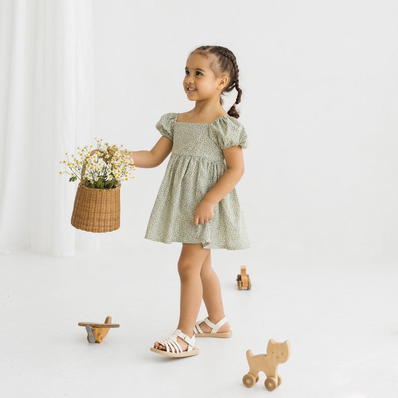 Puede incluir: Una ni&ntilde;a peque&ntilde;a con un vestido verde floral de mangas abullonadas sostiene una cesta de mimbre llena de flores blancas. Est&aacute; de pie sobre una superficie blanca con juguetes de madera esparcidos a su alrededor.