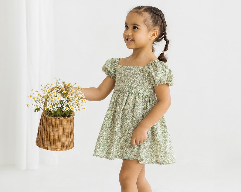 Puede incluir: Una ni&ntilde;a peque&ntilde;a con un vestido verde floral con mangas abullonadas y un escote cuadrado. Lleva una peque&ntilde;a cesta de mimbre llena de flores blancas.